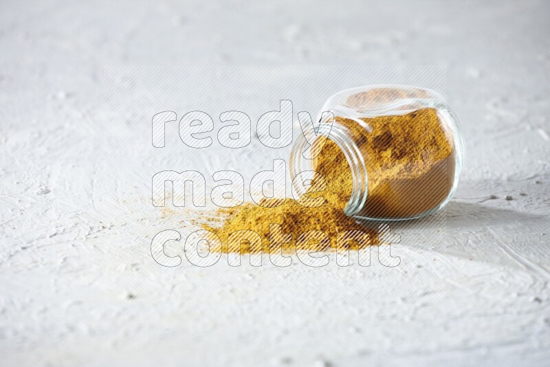 A flipped glass spice jar full of turmeric powder and powder fell out it on textured white flooring