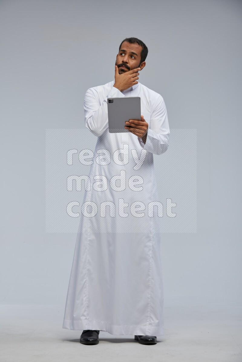 Saudi man wearing Thob standing working on tablet on Gray background