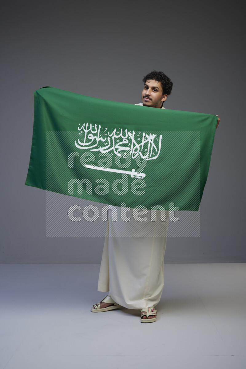 A Saudi man standing wearing thob holding big Saudi flag on gray background