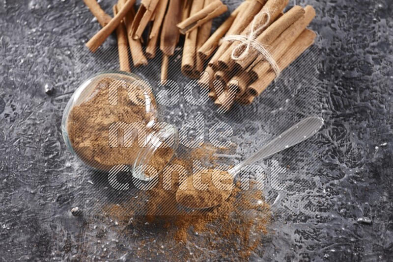 Herbal glass jar full cinnamon powder flipped and a metal spoon full of powder, cinnamon sticks stacked and bounded in the back on textured black background in different angles