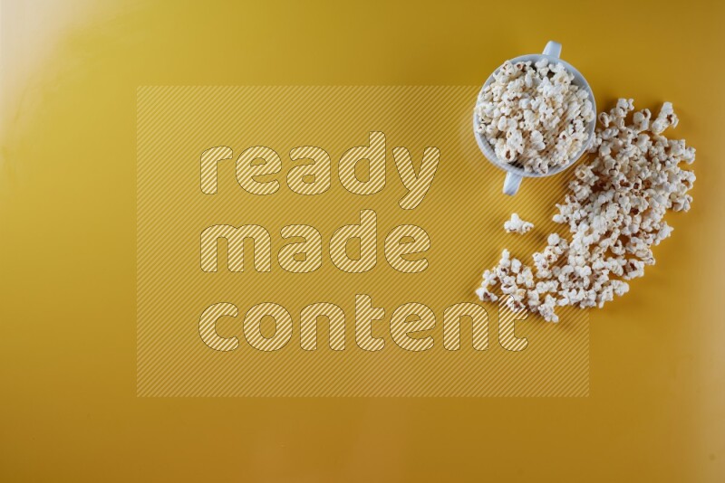 A white ceramic bowl full of popcorn with popcorn beside it on a yellow background in a top view shot
