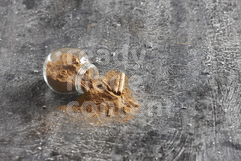 Flipped glass herbs jar full of cinnamon powder with cracked cinnamon sticks on a textured black background