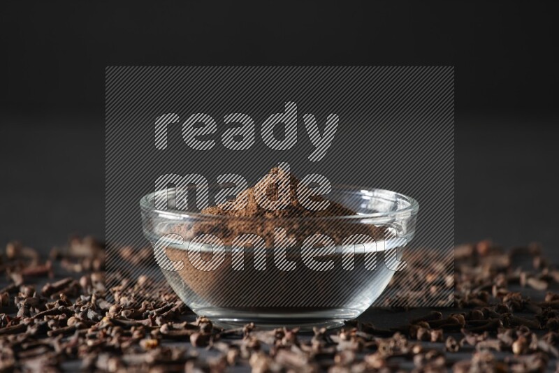 وعاء زجاجي ممتلئ ببودرة القرنفل مع حبوب القرنفل المتناثرة على أرضية سوداء