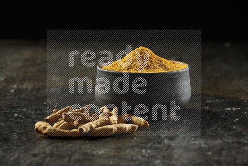 A black pottery bowl full of turmeric powder and dried turmeric whole fingers next of it on textured black flooring