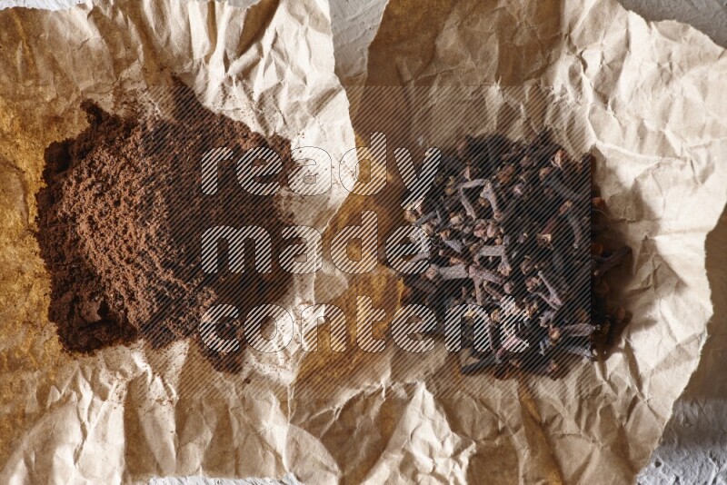 Cloves powder and cloves on 2 crumpled pieces of paper on a textured white flooring