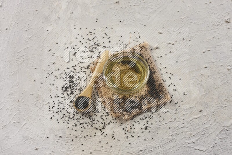 A glass bowl full of black seeds oil and wooden spoon full of black seeds with seeds spread on burlap fabric on a textured white flooring