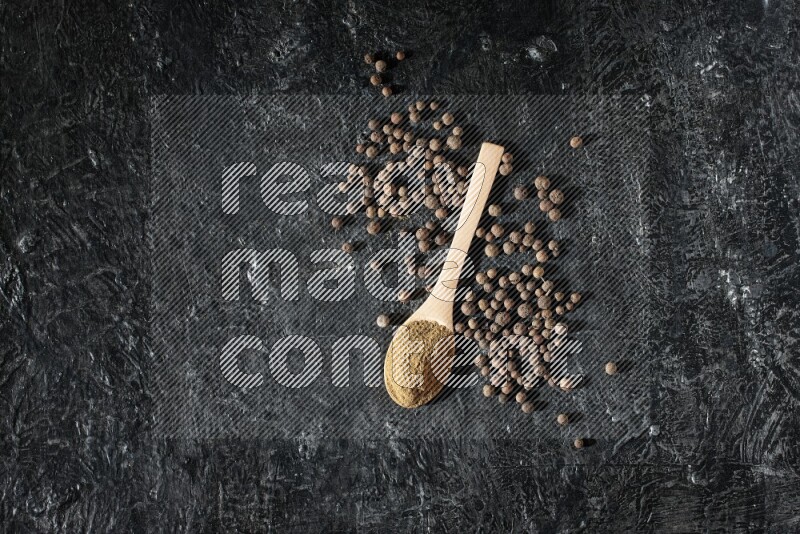 Wooden spoons full of all spice powder and allspice whole balls beside it on a textured black flooring