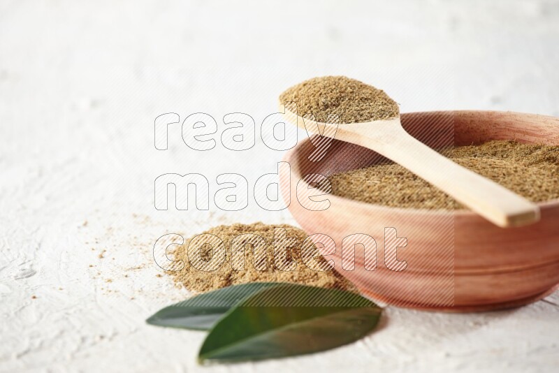 A wooden bowl and wooden spoon full of cumin powder on textured white flooring