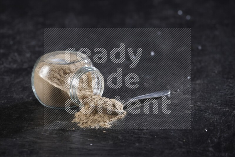 A glass spice jar full of garlic powder flipped and the powder came out with a filled metal spoon beside it on a textured black flooring