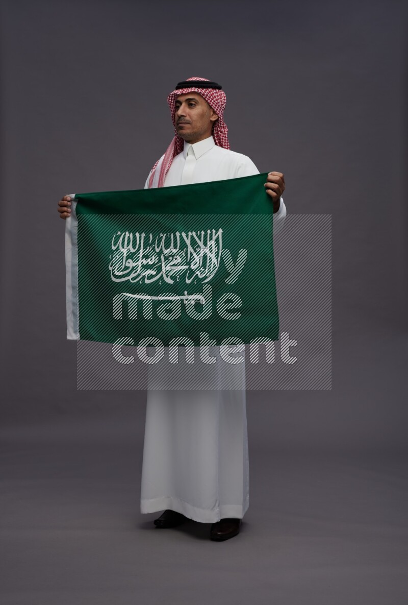 Saudi man wearing thob and shomag standing holding Saudi flag on gray background