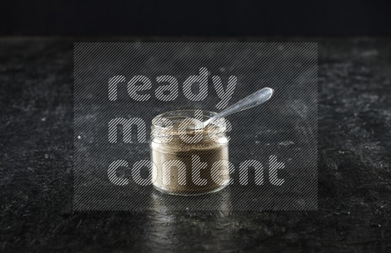 وعاء زجاجي للتوابل وملعقة معدنية ممتلئان ببودرة الهيل على خلفية سوداء