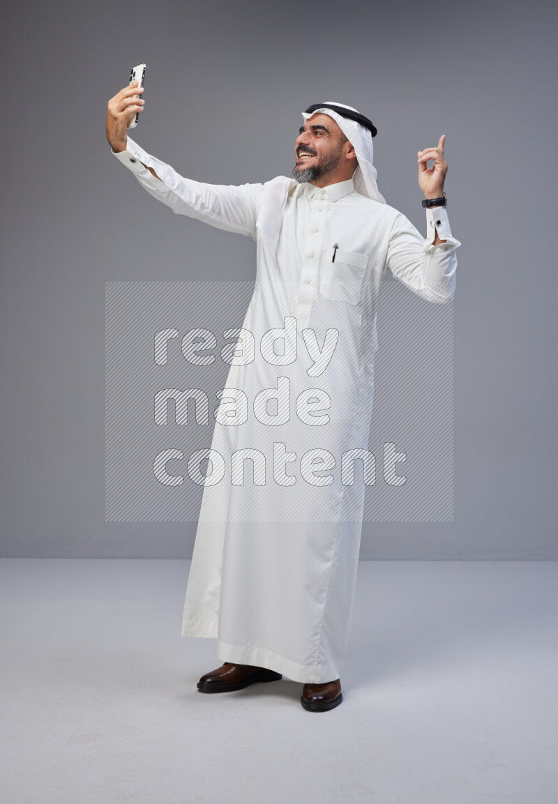 Saudi man Wearing Thob and white Shomag standing taking selfie on Gray background