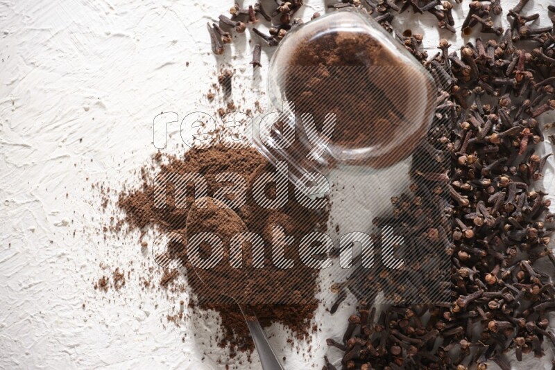 A flipped glass spice jar and a metal spoon full of cloves powder and powder came out of the jar with cloves spread on textured white flooring