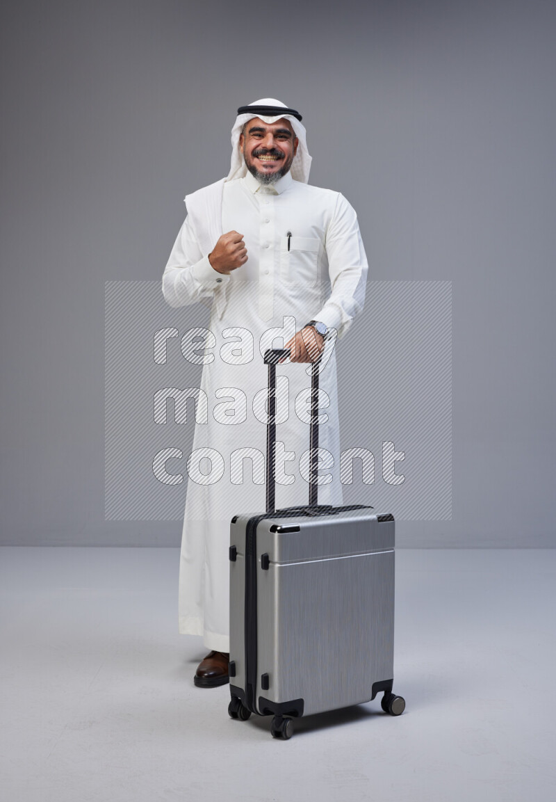Saudi man wearing Thob and white Shomag standing holding Travel bag on Gray background