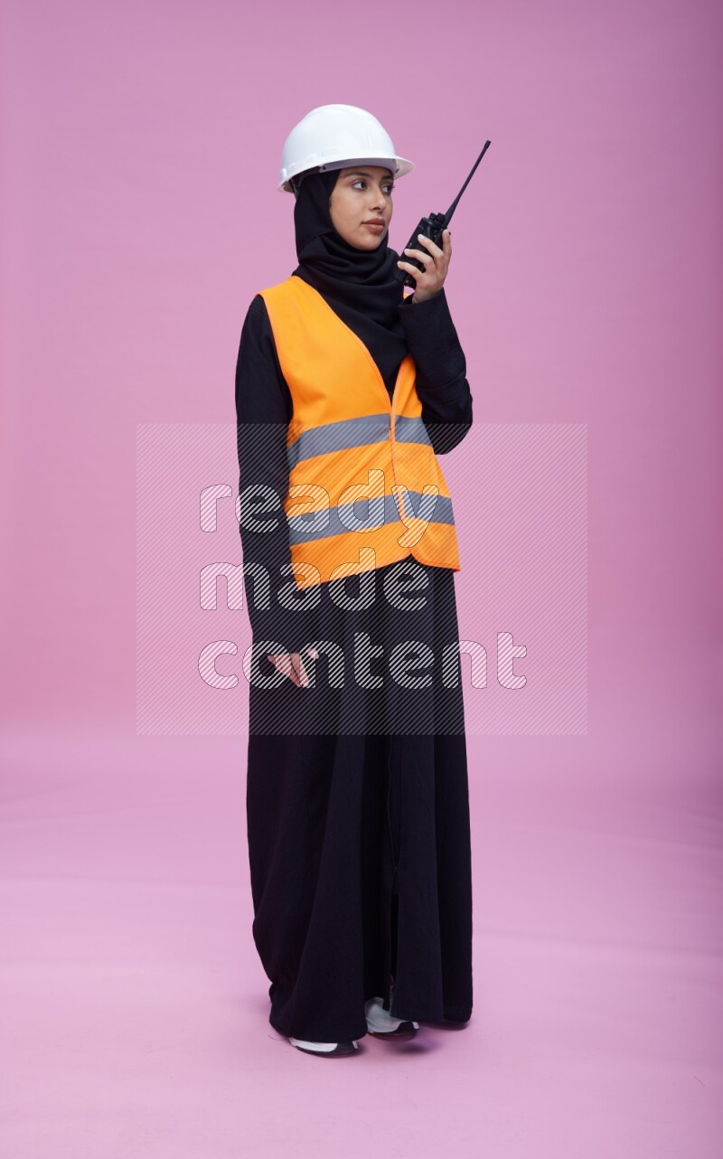 Saudi woman wearing Abaya with engineer vest and helmet standing holding walkie-talkie on pink background