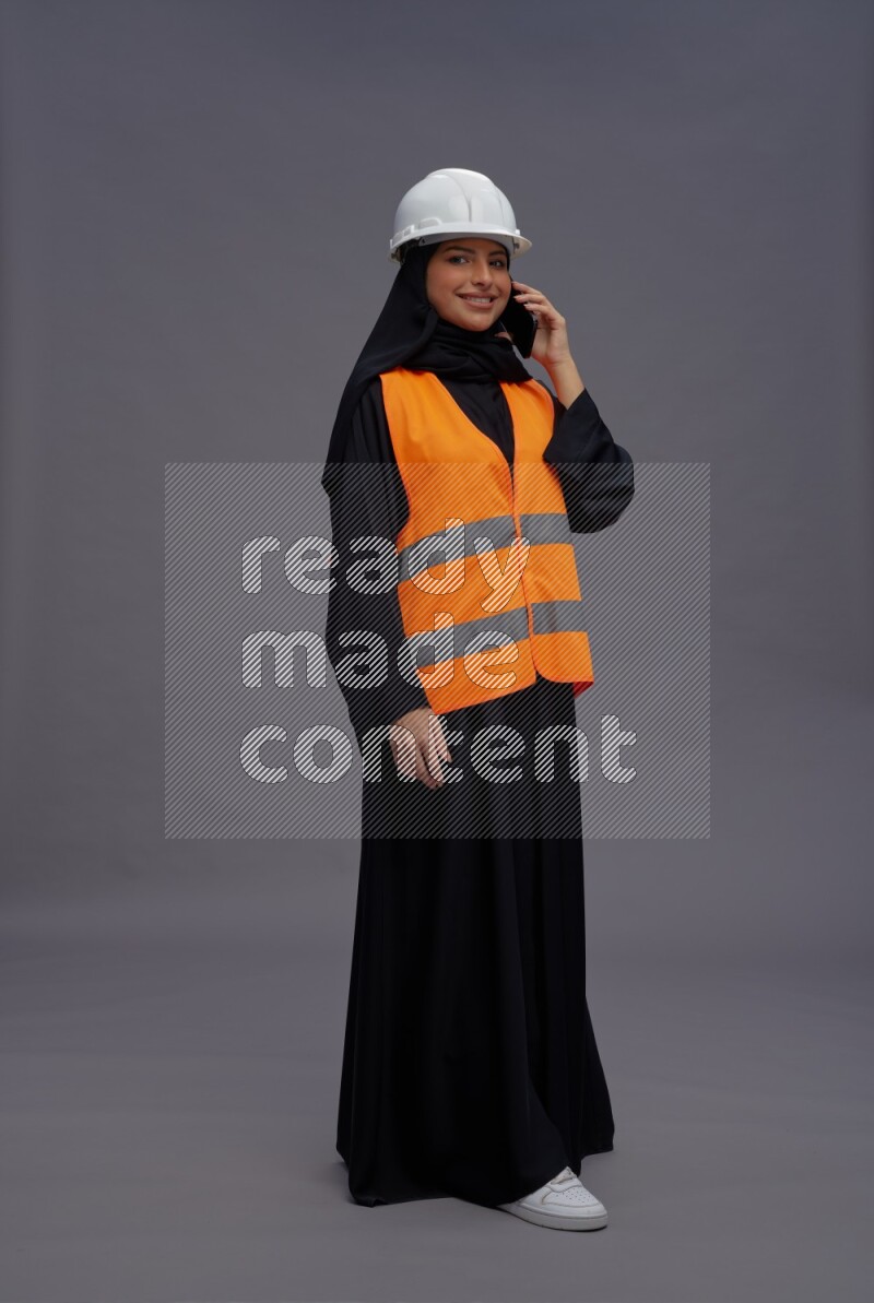 Saudi woman wearing Abaya with engineer vest standing talking on phone on gray background