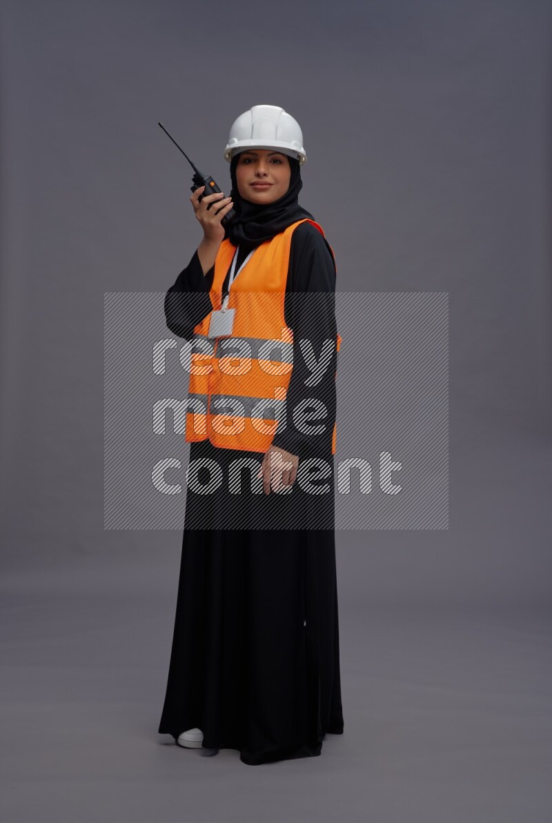 Saudi woman wearing Abaya with engineer vest with neck strap employee badge standing holding walkie-talkie on gray background