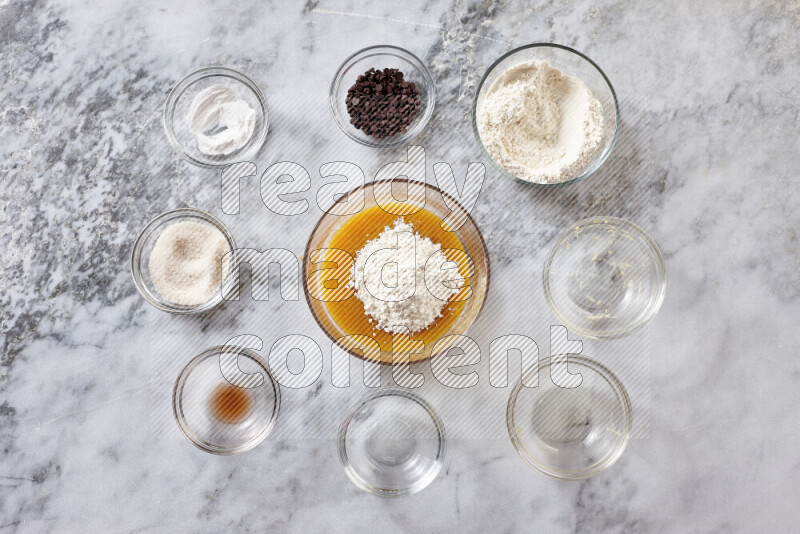 كوكيز خطوة بخطوة بمكوناتها، الدقيق، الزبدة، السكر البني، البيض، الفانيليا، السكر الأبيض، رقائق الشوكولاتة وصودا الخبز على خلفية رخام رمادي