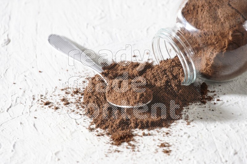 وعاء زجاجي مقلوب وملعقة معدنية ممتلئان ببودرة القرنفل وتناثرت البودرة من الوعاء مع حبوب القرنفل المنتشرة على أرضية بيضاء