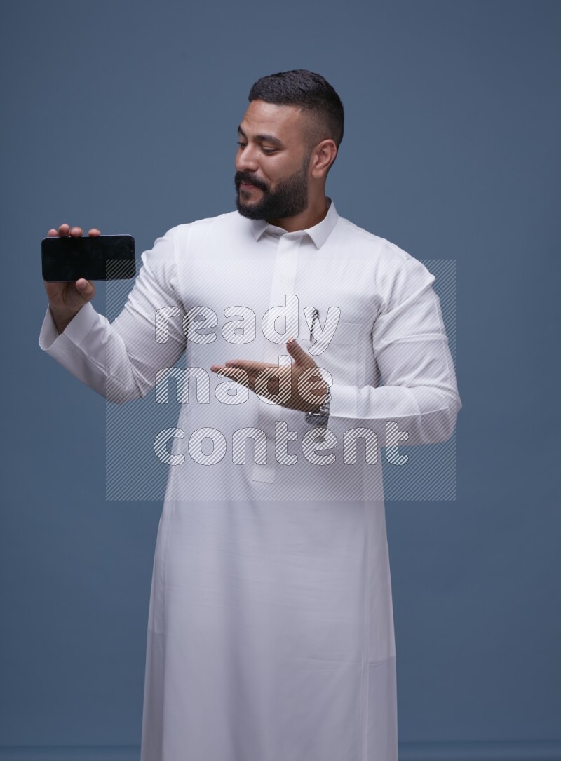 A man Showing his phone in Blue Background wearing Saudi Thob