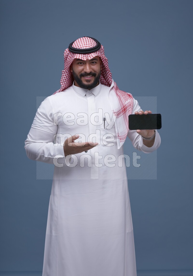A man Showing his smartphone Screen on Blue Background wearing Saudi Thob