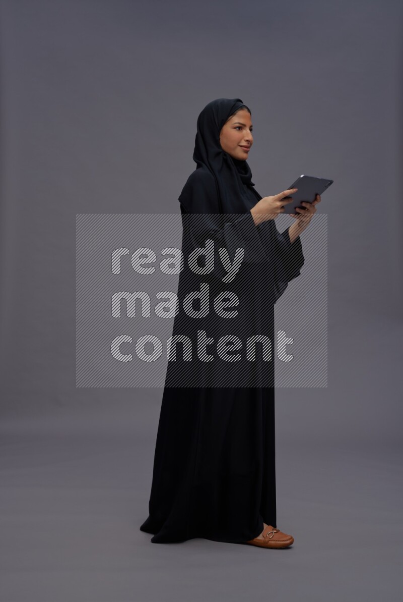 Saudi woman wearing Abaya standing working on tablet on gray background