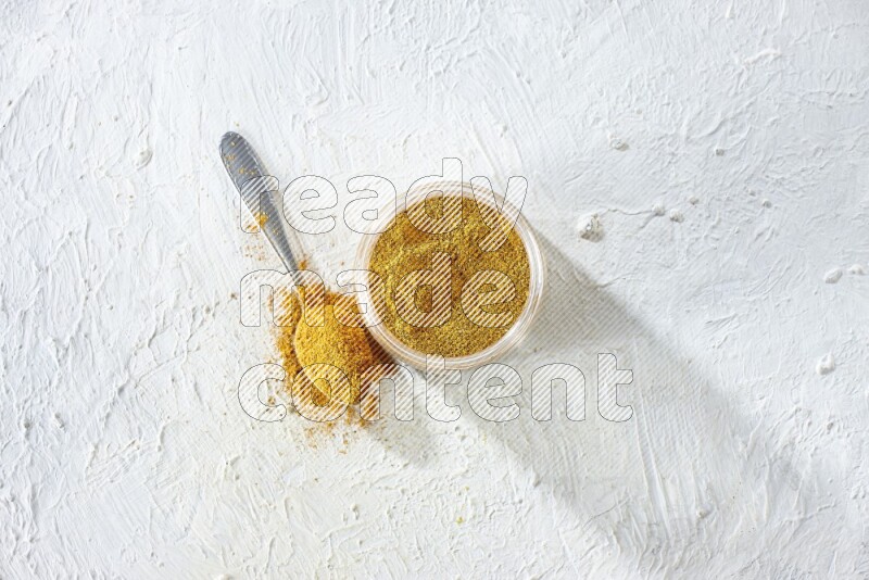 A glass jar and a metal spoon full of turmeric powder on a textured white flooring