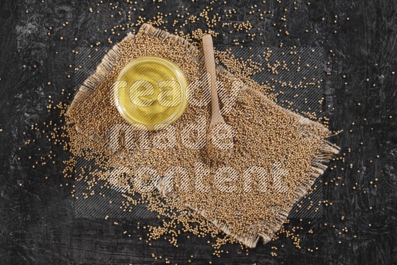 A glass jar full of mustard paste set on a burlap piece with a wooden spoon full of mustard seeds on a textured black flooring