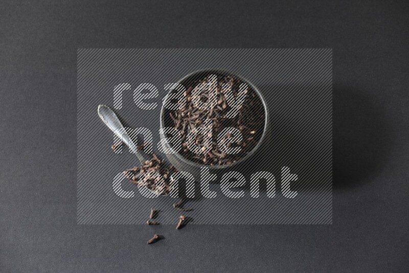 وعاء فخاري أسود ممتلئ بحبوب القرنفل مع ملعقة معدنية بجانبه علي خلفية سوداء