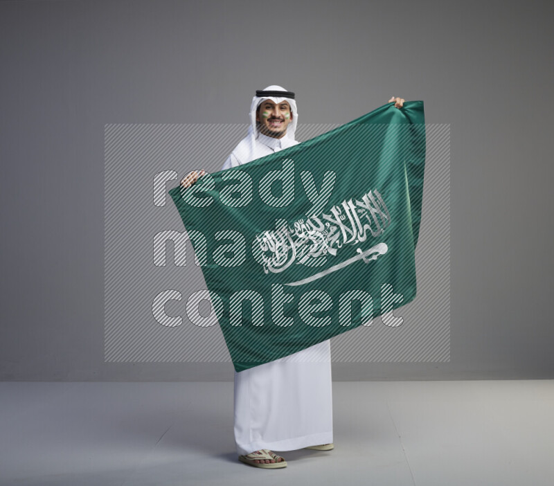 A Saudi man standing wearing thob and white shomag with face painting holding big Saudi flag on gray background