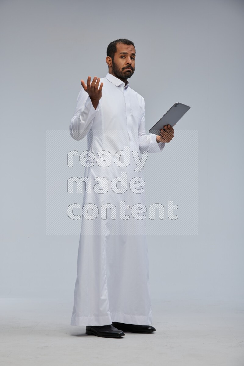 Saudi man wearing Thob standing working on tablet on Gray background