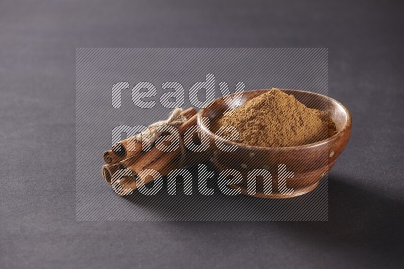 Cinnamon sticks stacked and bounded beside a wooden bowl full of cinnamon powder and a wooden spoon full of powder on black background