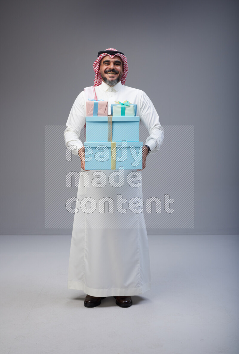 Saudi man Wearing Thob and red Shomag standing holding gift box on Gray background