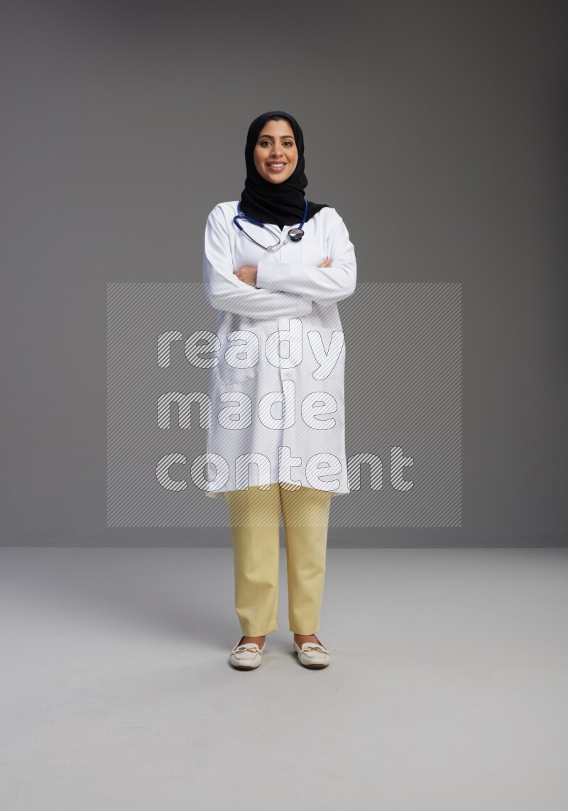 Saudi woman wearing lab coat with stethoscope standing with crossed arms on Gray background
