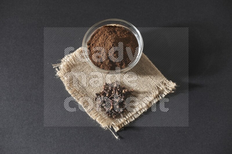 A glass bowl full of cloves powder with cloves grains on a burlap piece on a black flooring