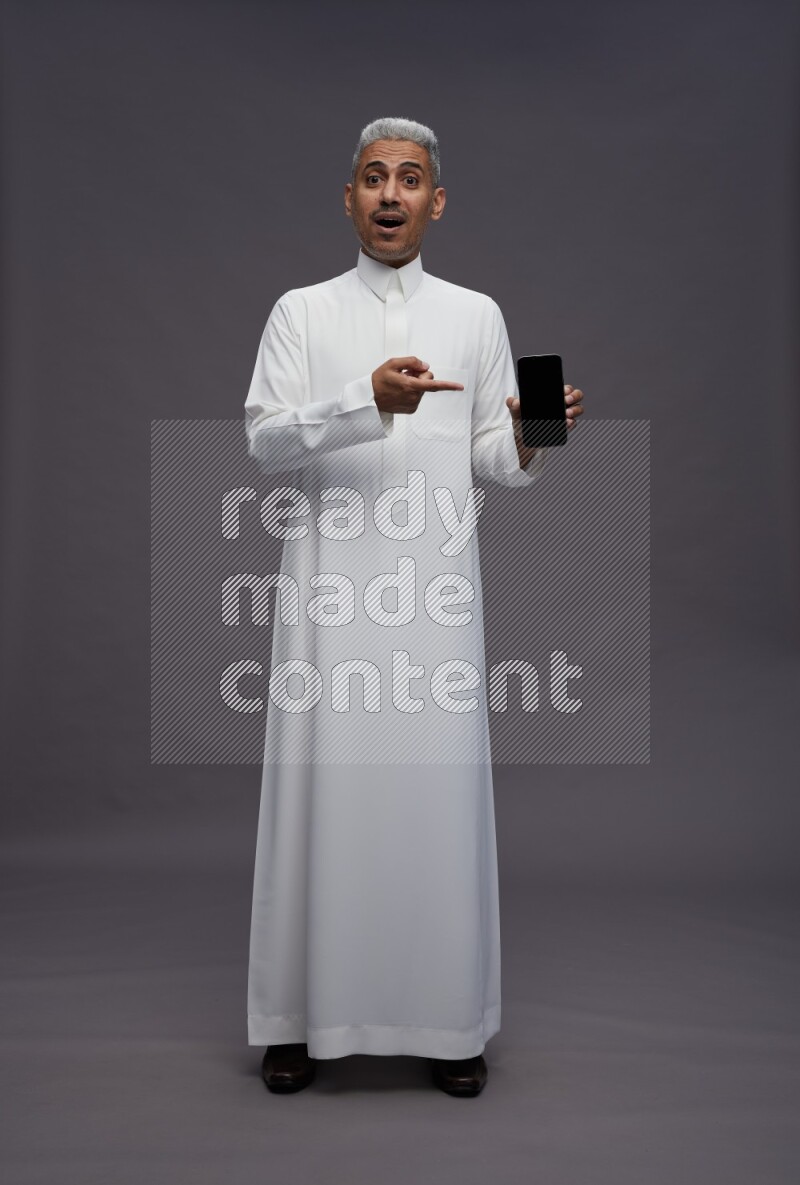 Saudi man wearing thob standing showing phone to camera on gray background