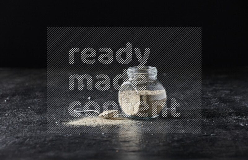 Herbal glass jar and metal spoon full of white pepper powder on textured black flooring