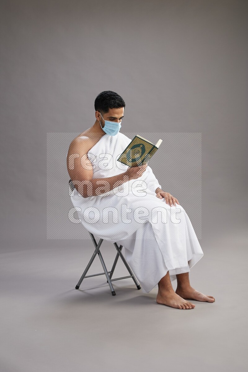 A man wearing Ehram with face mask sitting on chair reading quran on gray background