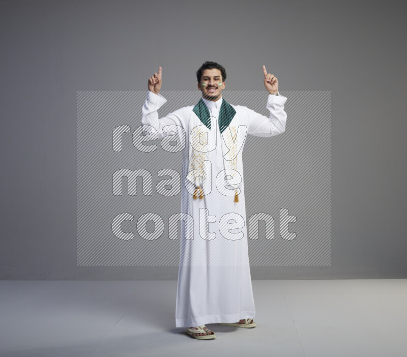 A Saudi man standing wearing thob and Saudi flag scarf with face painting on gray background