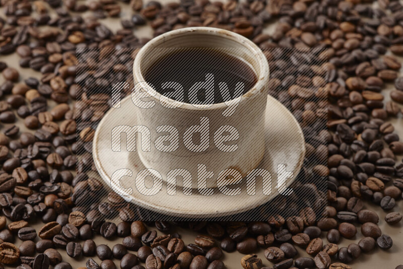 A beige pottery cup of coffee surrounded by roasted coffee beans on beige background