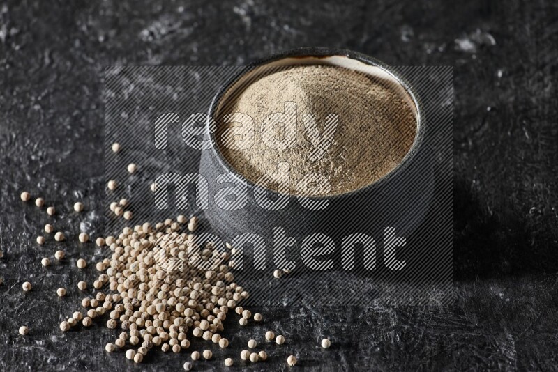 وعاء فخار أسود مملوء ببودرة الفلفل الأبيض مع حبات الفلفل الأبيض على أرضية سوداء