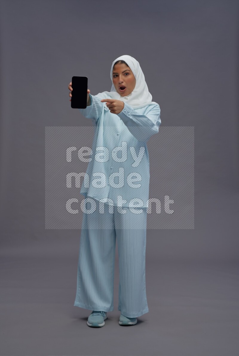 Saudi woman wearing hijab clothes standing showing phone to camera on gray background