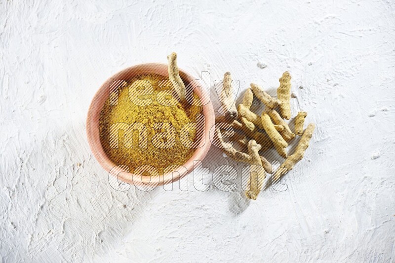 A wooden bowl full of turmeric powder and dried turmeric whole fingers beside it on textured white flooring