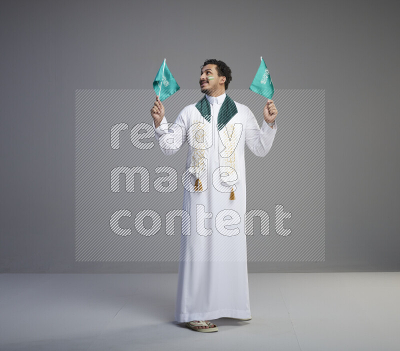 A Saudi man standing wearing thob and saudi flag scarf with face painting holding small Saudi flag on gray background