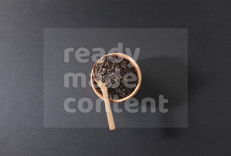 A wooden bowl and a wooden spoon full of cloves on a black flooring