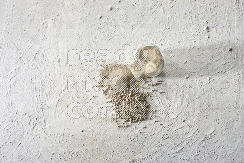 وعاء زجاجي عشبي مقلوب ممتلئ ببودرة الفلفل الأبيض مع بودرة وحبوب متناثرة على أرضية بيضاء