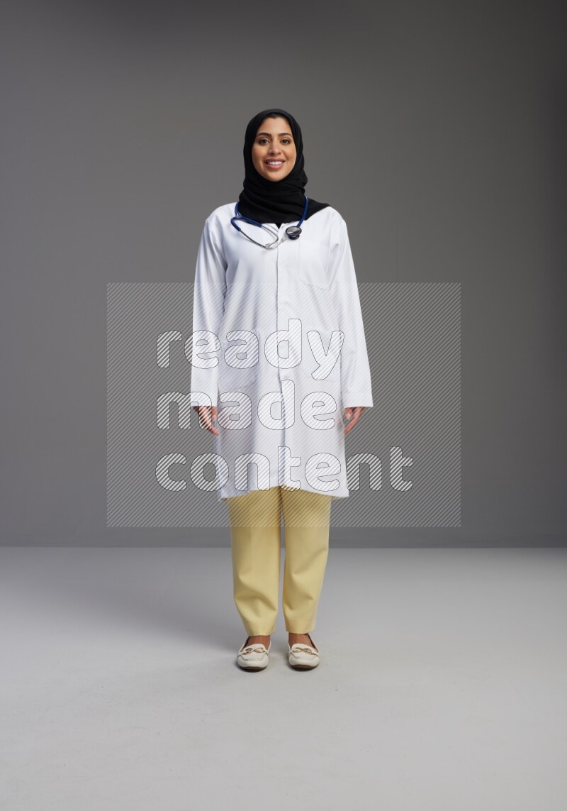 Saudi woman wearing lab coat with stethoscope standing interacting with the camera on Gray background