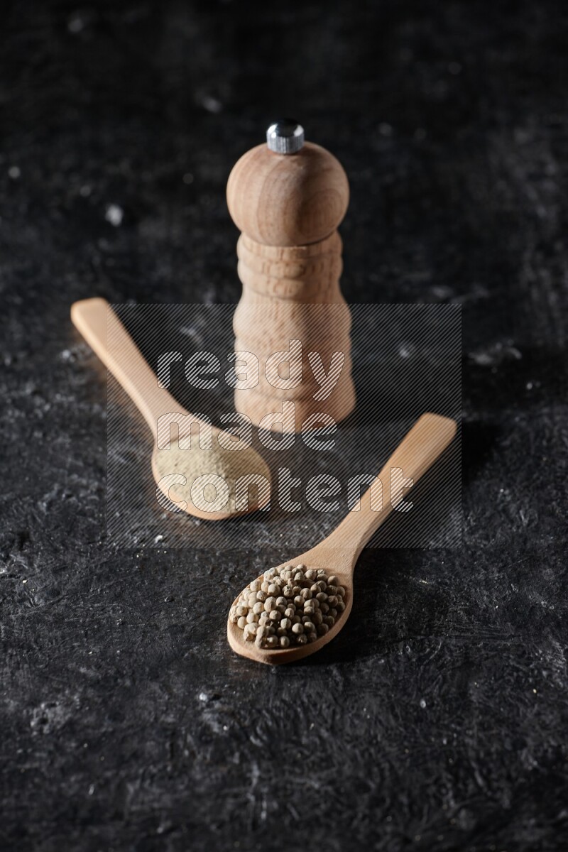 2 wooden spoons one full of white pepper powder and the other with pepper beads and a wooden pepper mill on textured black flooring