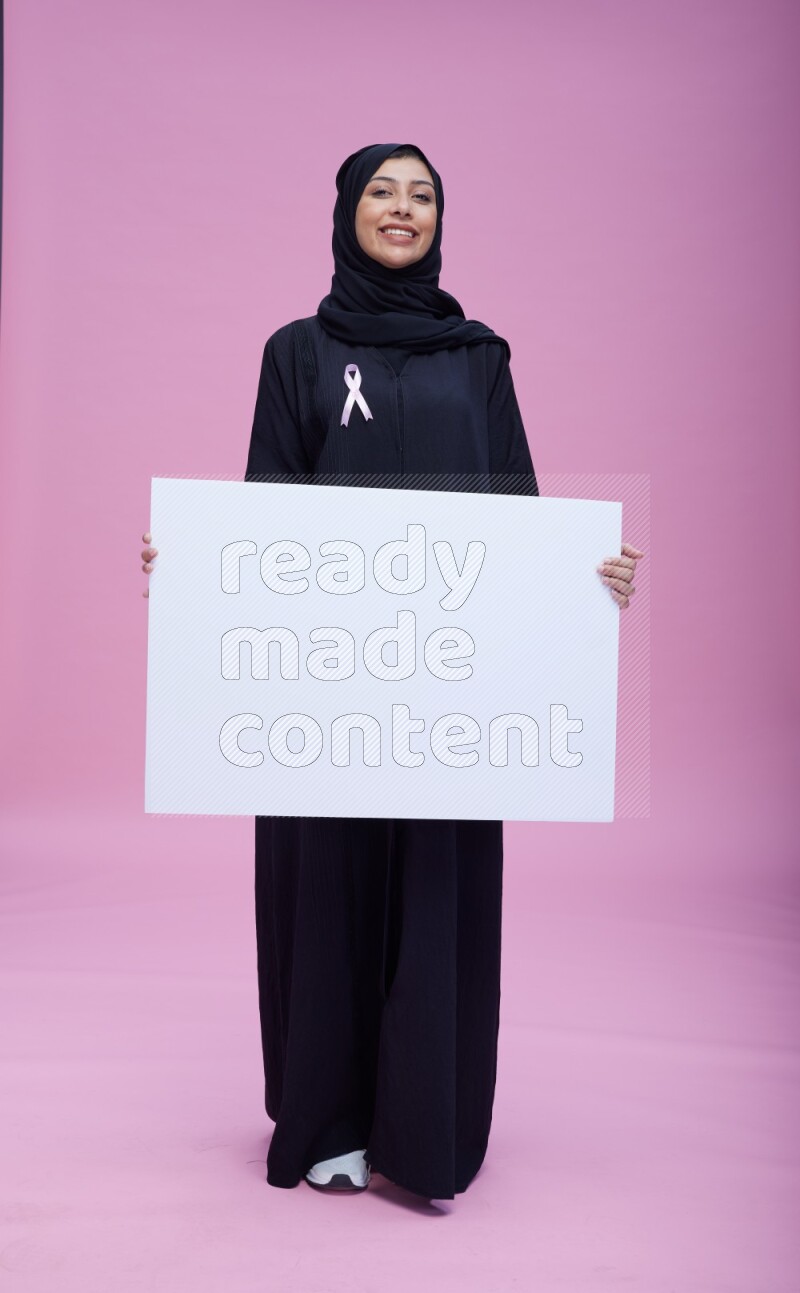 Saudi woman wearing pink ribbon on Abaya standing holding board on pink background