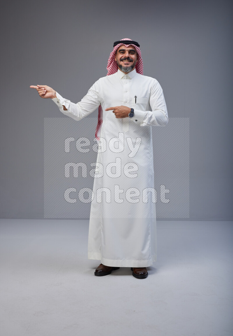 Saudi man Wearing Thob and red white Shomag standing interacting with the camera on Gray background